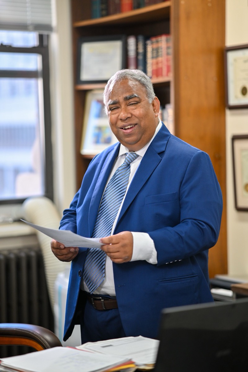 罗曼大律师 Meer Md. Mizanur Rahman 在办公室内站立阅读文件，身后为执业证书墙与法律藏书